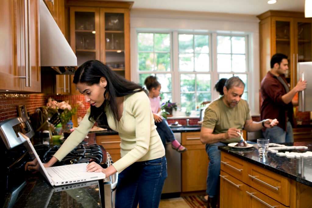 Woman looks at laptop while multigenerational family moves around kitchen in background