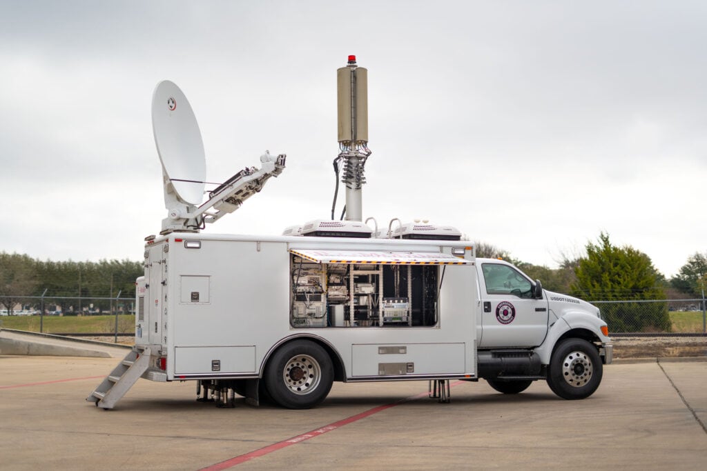 Large truck with mounted satellite dish and antenna tower. Vehicle is branded with the T-Mobile Emergency Management logo and has an open window to display the interior equipment.