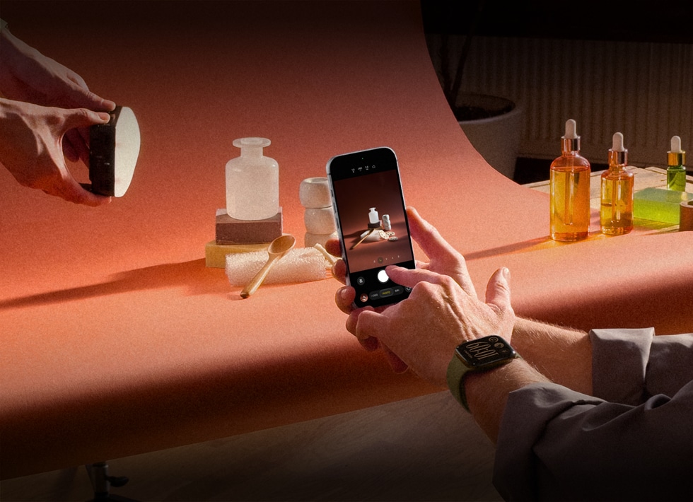 En una sesión fotográfica de productos, unas manos, con un dispositivo Apple Watch en la muñeca izquierda, sostienen un iPhone 18 Pro, mientras toman fotos de los productos, otras manos iluminan el producto
