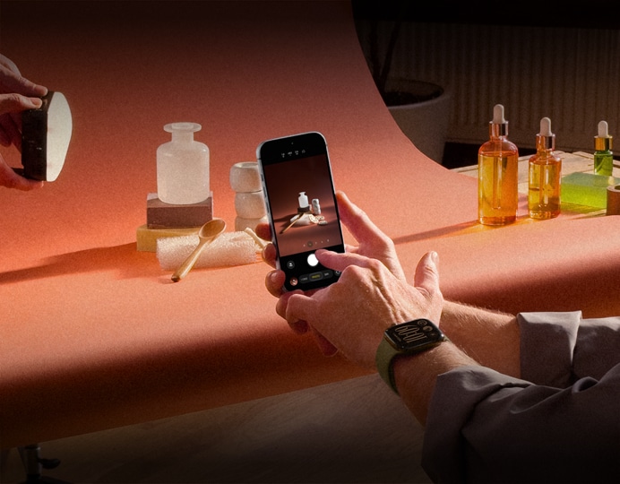 En una sesión fotográfica de productos, unas manos, con un dispositivo Apple Watch en la muñeca izquierda, sostienen un iPhone 18 Pro, mientras toman fotos de los productos, otras manos iluminan el producto