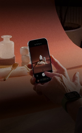 En una sesión fotográfica de productos, unas manos, con un dispositivo Apple Watch en la muñeca izquierda, sostienen un iPhone 18 Pro, mientras toman fotos de los productos, otras manos iluminan el producto