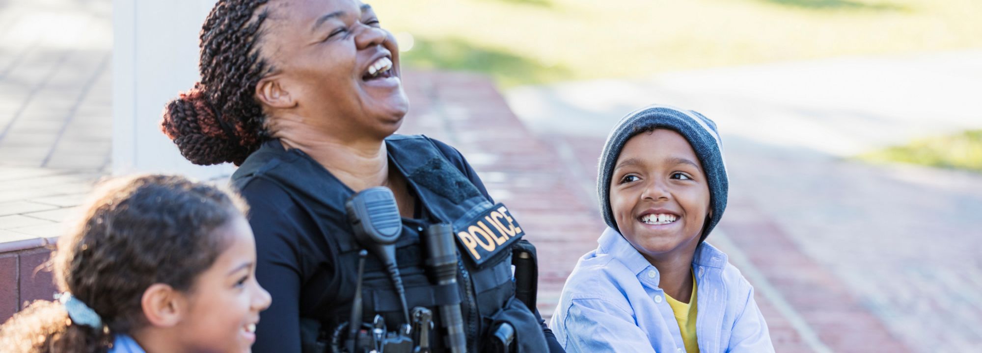 Un policía sonriente sentado al aire libre con dos niños, todos ríen juntos en la acera.