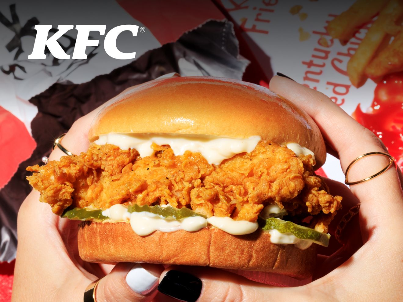 Unas manos sostienen un sándwich de pollo de KFC sobre envoltorios de KFC y papas fritas