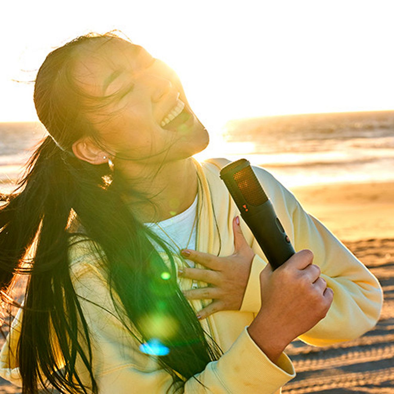 Una mujer canta en la playa con la puesta de sol a sus espaldas usando la JBL PartyBox On-the-Go 2.