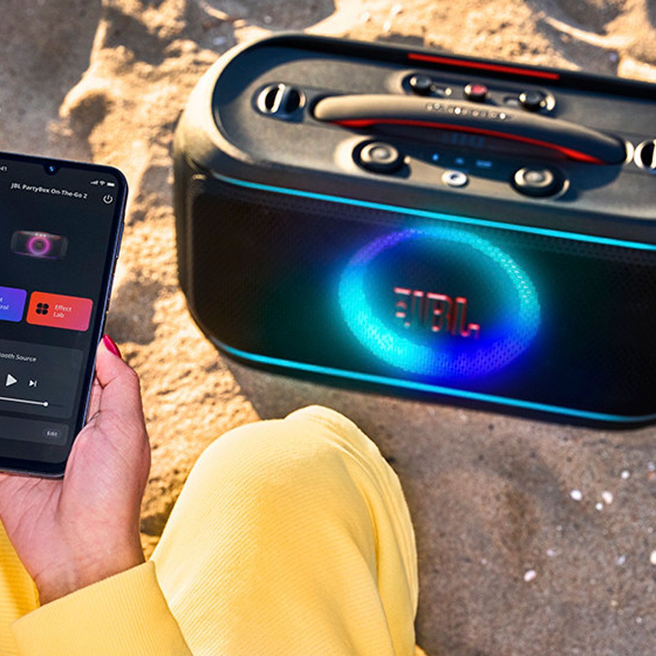 Una mujer en la playa usa la app JBL PartyBox en su teléfono para reproducir música en la JBL Partybox On-the-Go 2.