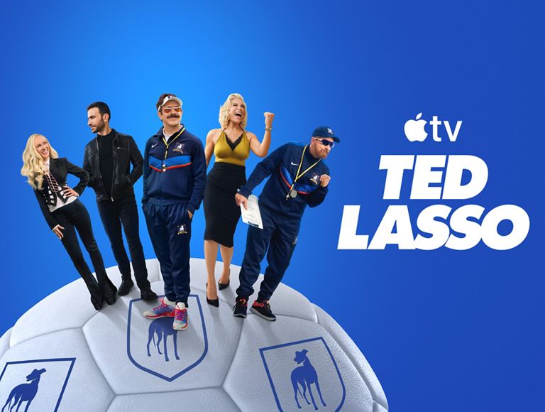 Imagen promocional de Apple TV+ en la que aparece el reparto de Ted Lasso de pie sobre un balón de fútbol con un fondo azul.