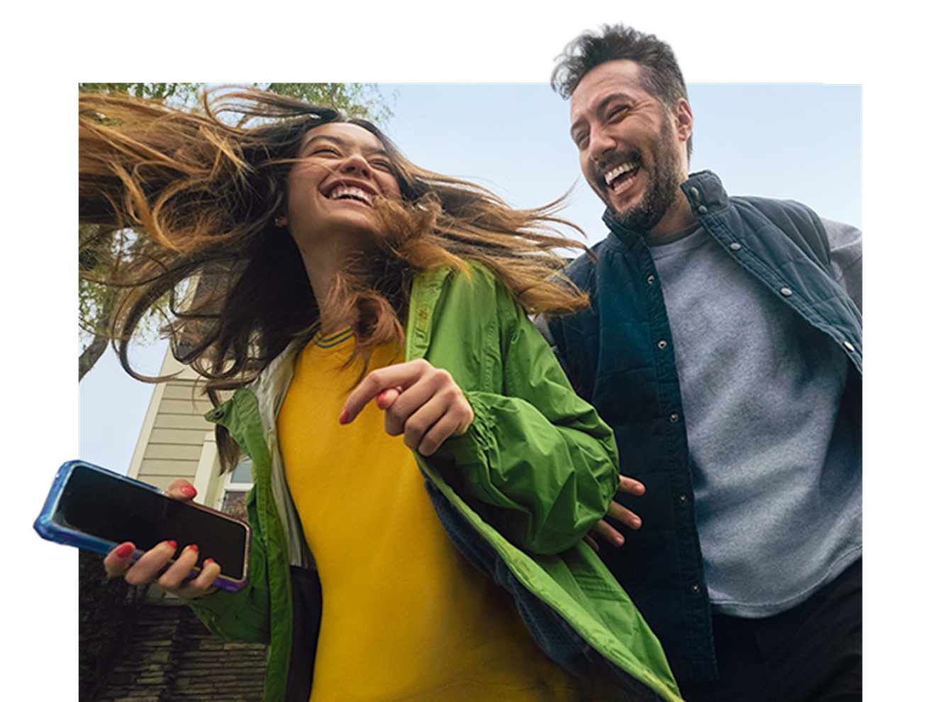 Un hombre y una mujer sonrientes con chaquetas se mueven fuera de una casa. Ella sostiene un teléfono azul en la mano.