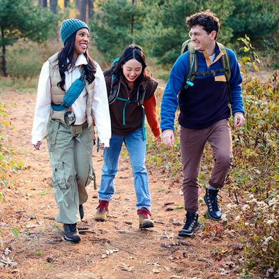 Dos mujeres y un hombre sonríen mientras pasean juntos por el bosque con su JBL Clip five.