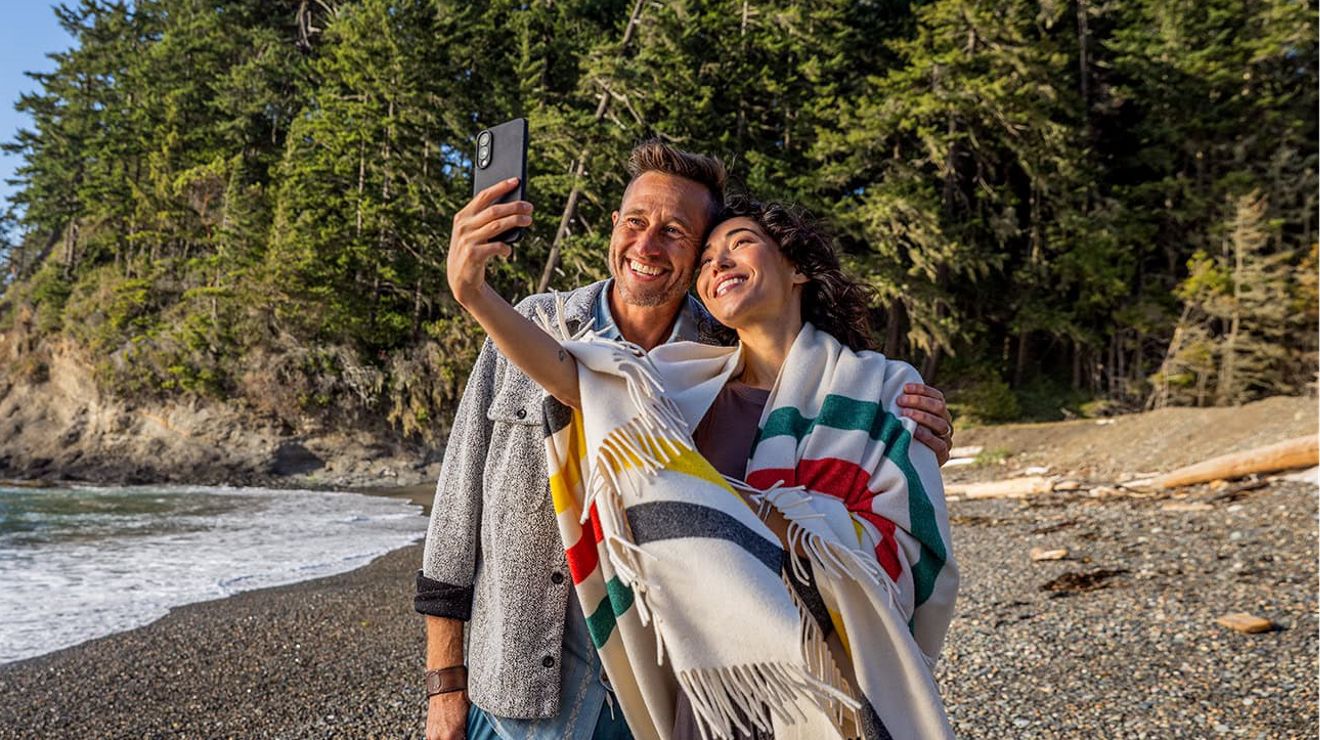 Bajo un hermoso cielo azul, una mujer y un hombre posan para un selfie en una playa de guijarros.