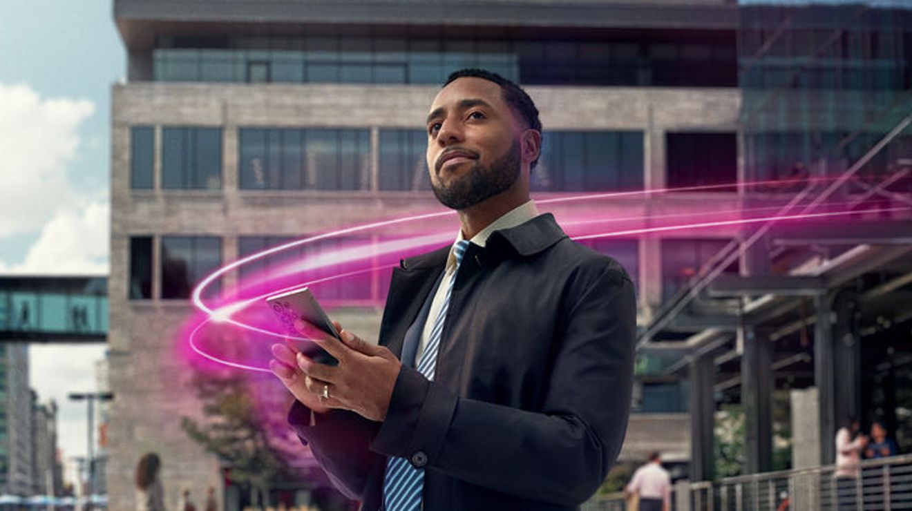 Un hombre vestido de manera profesional parado fuera de un edificio de oficinas moderno mira su smartphone mientras un haz de luz rosa brillante gira a su alrededor, sugiriendo conectividad digital o tecnología móvil.