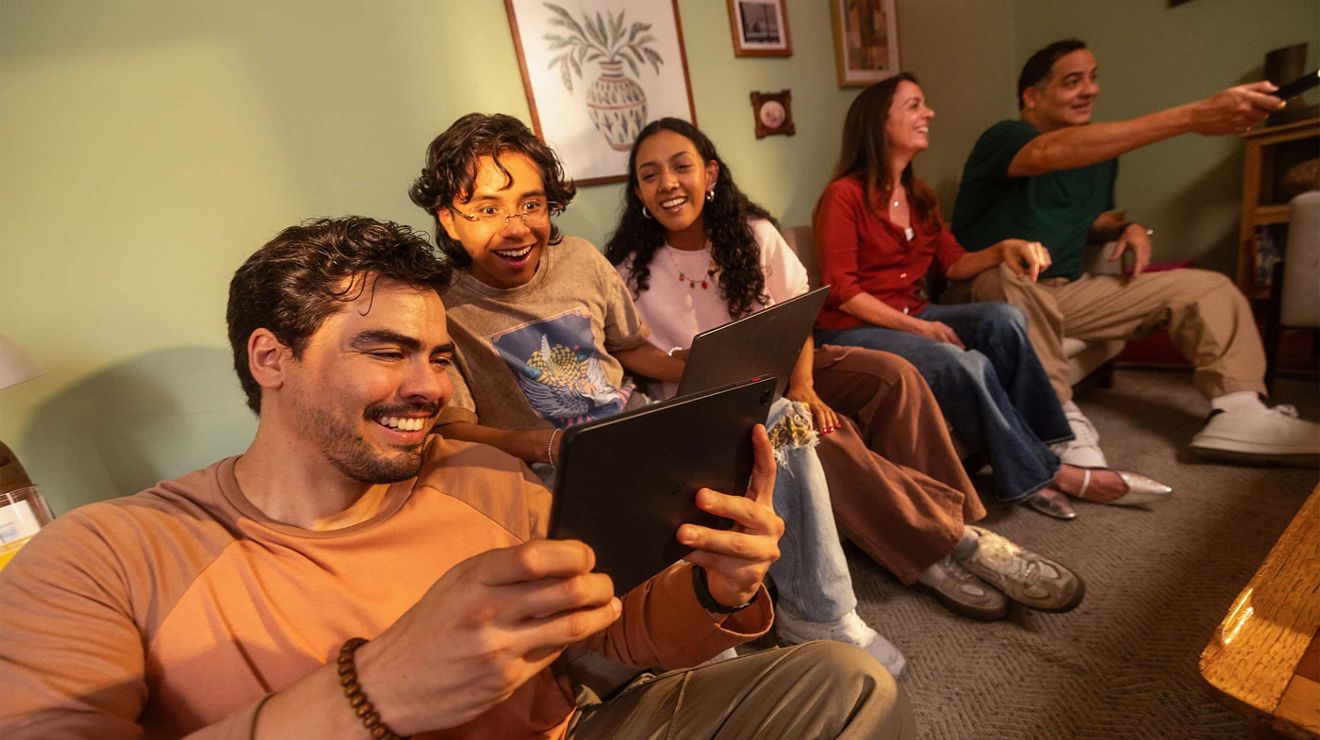 Un grupo de amigos reunidos en una sala de estar, usando smartphones y mirando una pantalla mientras están sentados en el sofá y en el suelo.