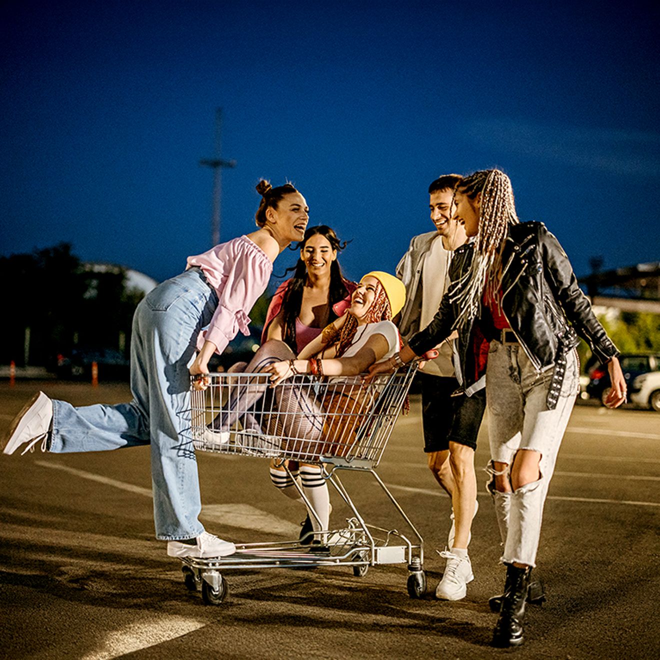 Un grupo de amigos se divierte al atardecer, empujando un carrito de compras por un estacionamiento vacío con gran energía.