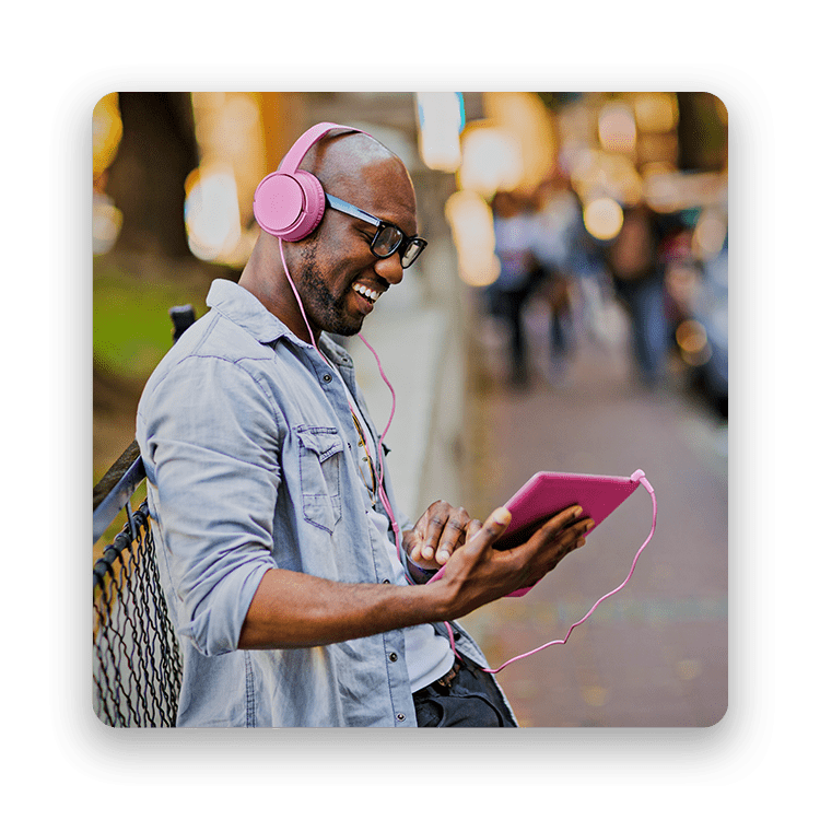 Hombre sonriente sostiene una tablet color magenta mientras escucha algo en sus audífonos magenta en la vereda