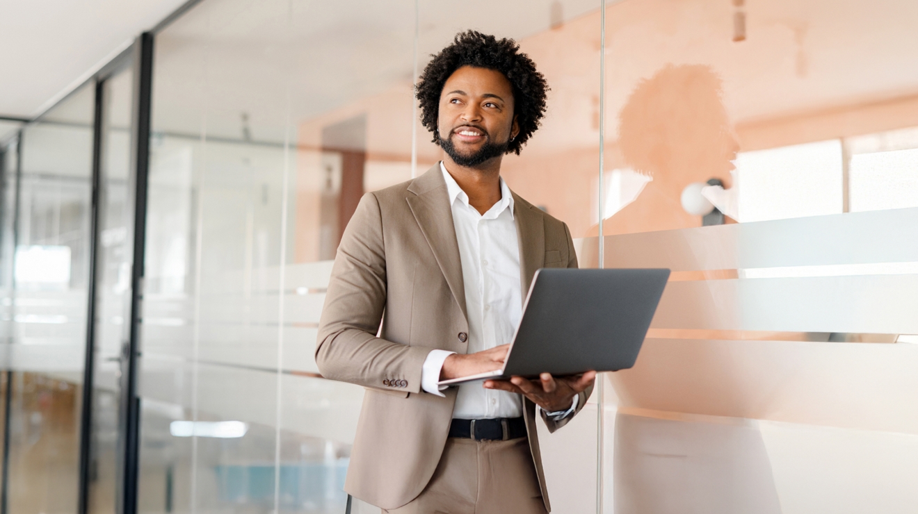 Un profesional con una laptop en la mano se detiene frente a una sala de conferencias con paredes de vidrio.