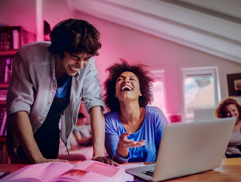 Dos amigos riendo mientras hacen tareas escolares en una laptop.