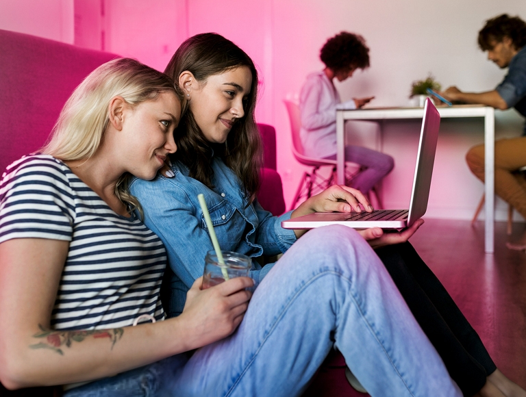 Dos amigas utilizan una laptop sentadas en el suelo detrás de un sofá.