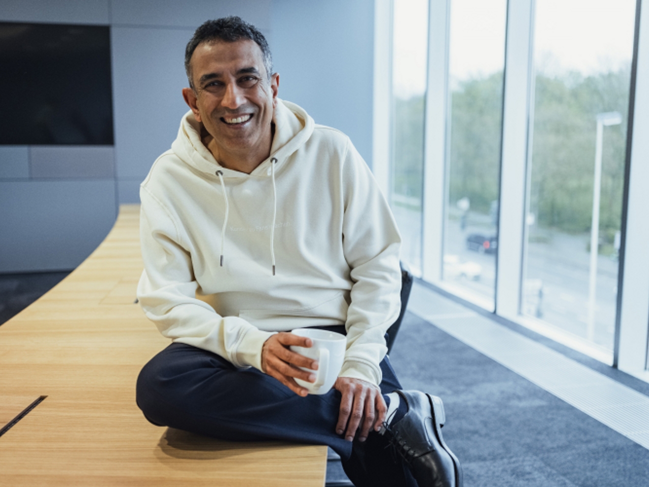 Srini sonríe mientras sostiene una taza de café y se apoya tranquilamente en una mesa.
