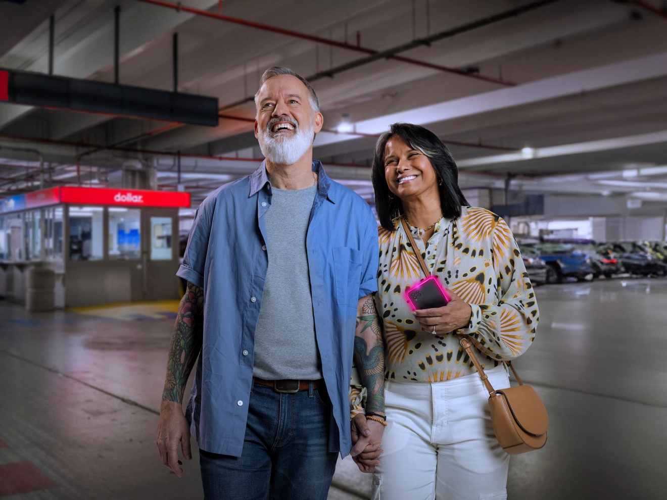 Pareja sonriendo en una tienda Dollar Car Rental; la mujer lleva en la mano un teléfono móvil con contorno magenta.