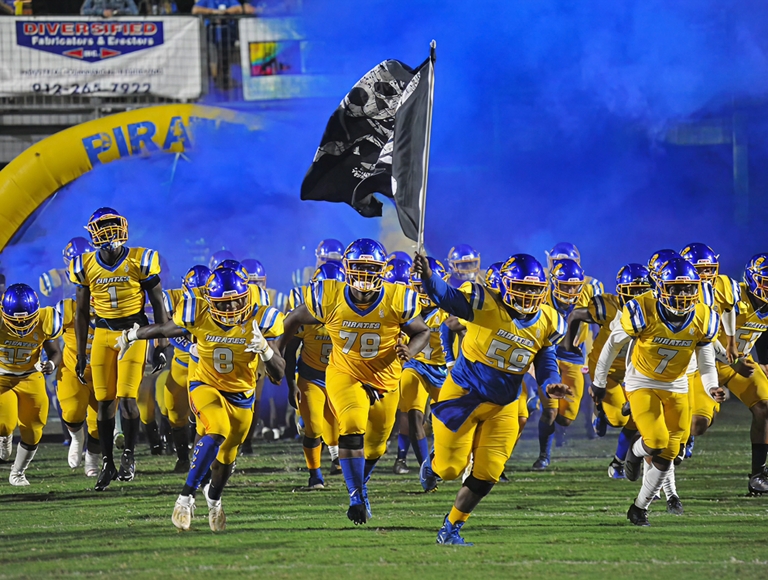 El equipo de fútbol de Brunswick High School​​​​​​​, con uniformes de color azul y amarillo, ingresa al campo haciendo flamear una bandera pirata.