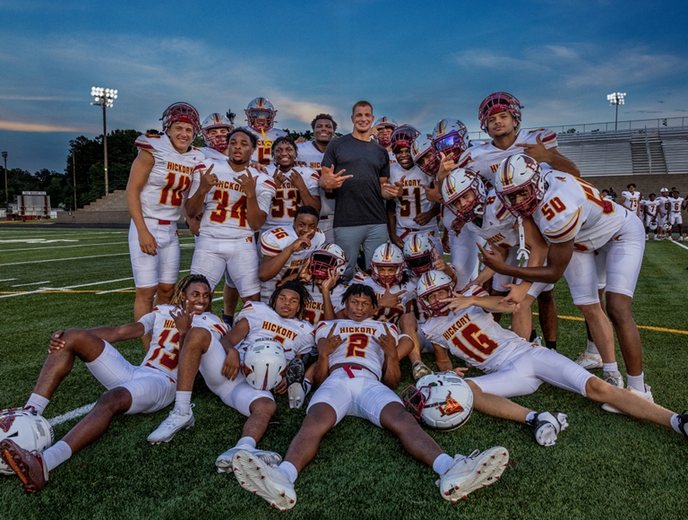 El equipo de fútbol de Hickory High School, con uniformes de color blanco, rojo y dorado, posa en grupo en un campo de fútbol al aire libre.​​​​​​​