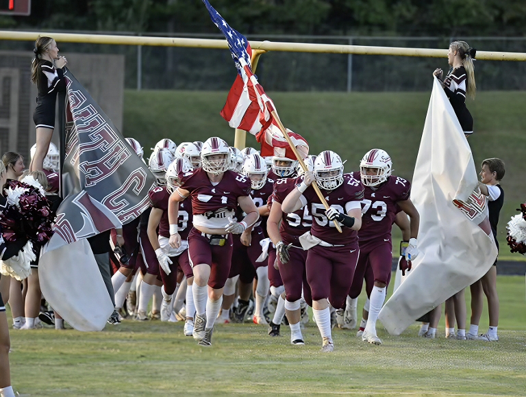 El equipo de fútbol de South Caldwell High School, con uniformes de color granate y blanco, ingresa al campo haciendo flamear varias banderas.