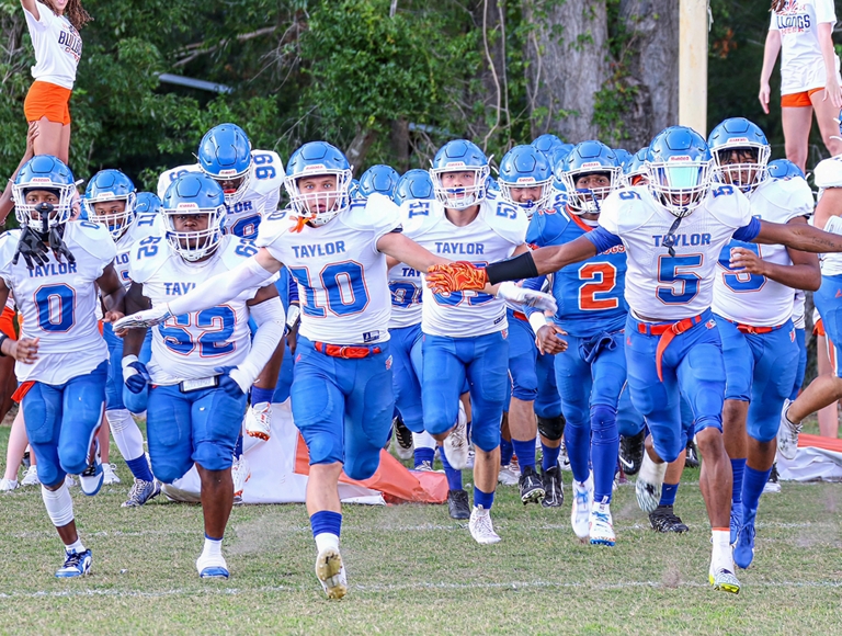 El equipo de fútbol de Taylor County High School, con uniformes de color naranja y azul, corre en el campo antes de un partido.