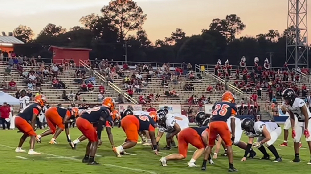 El equipo de fútbol de Escambia High School, con uniformes azul marino y naranja, entra al campo.