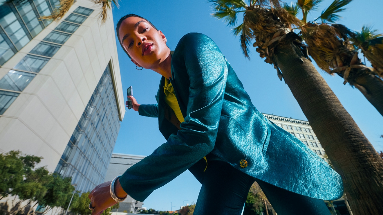 Mujer vestida con un traje azul mira hacia abajo mientras sonríe rodeada por edificios y palmeras.