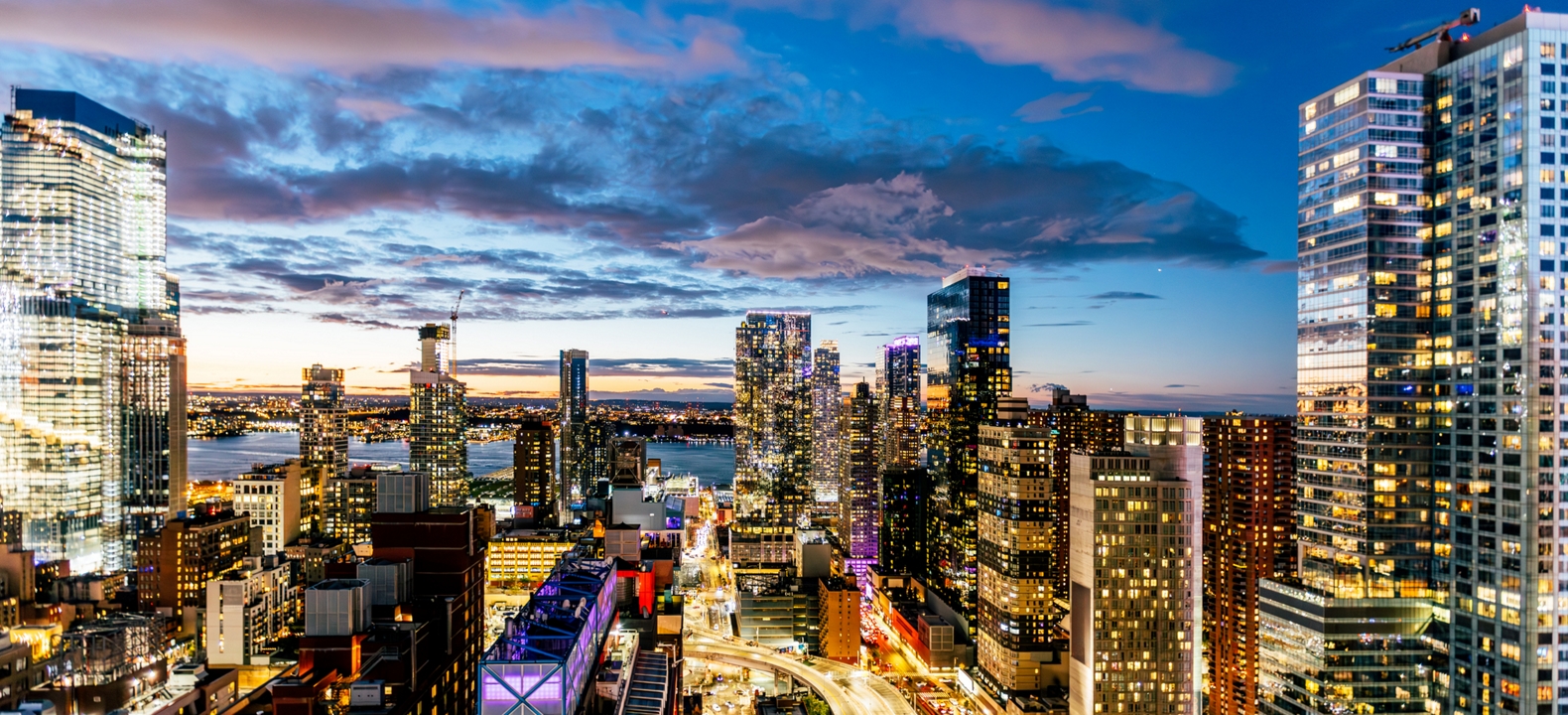 Un paisaje urbano al atardecer, con calles bulliciosas iluminadas por las luces de los autos y rascacielos que se recortan contra el cielo.