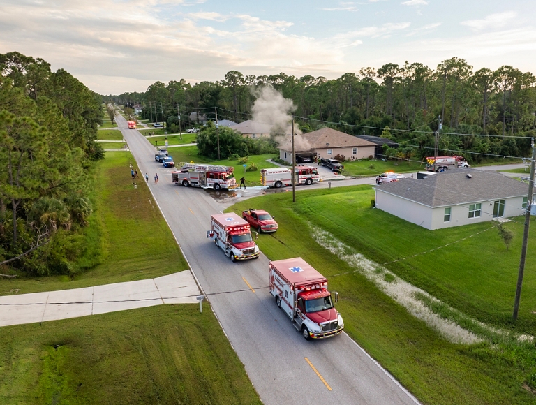 Una vista aérea muestra varios vehículos de emergencia, incluidos camiones de bomberos y ambulancias, reunidos en una calle suburbana donde sale humo de una casa.