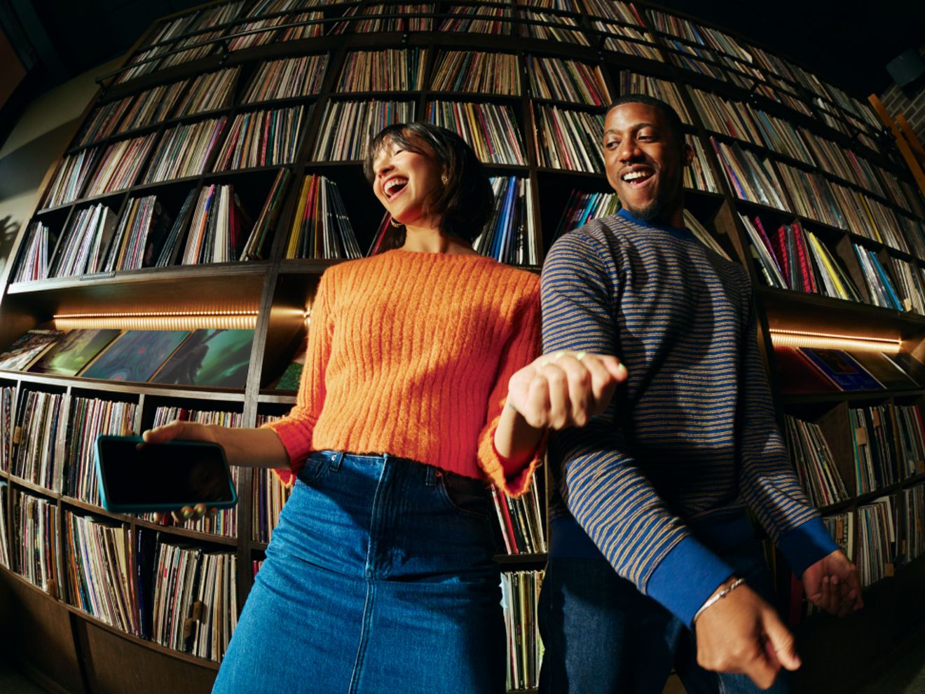 Una mujer con un suéter naranja y un hombre con un suéter a rayas sonríen y bailan frente a una pared de discos de vinilo en estantes. Ella sostiene un teléfono azul en la mano.