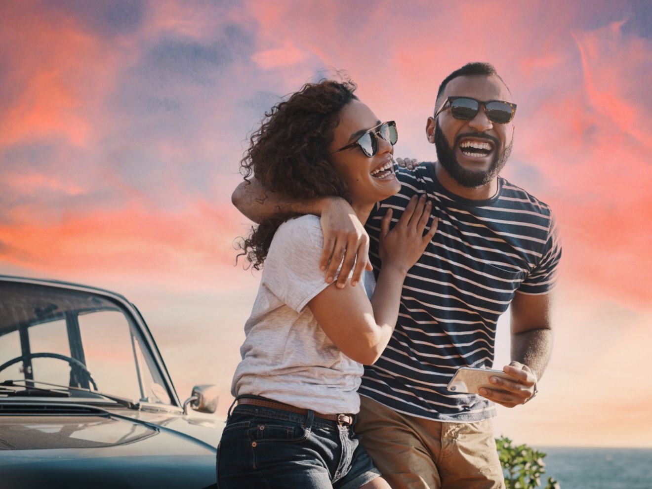 Una pareja se ríe mientras está sentada en su coche estacionado. Es de noche y la puesta de sol queda a sus espaldas.