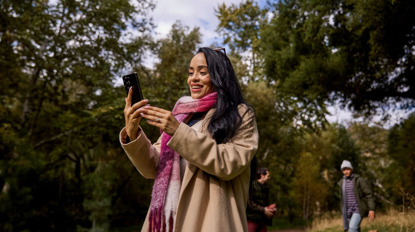 Una mujer con una bufanda a cuadros magenta y blancos está de pie al aire libre, rodeada de árboles, sonriendo a su teléfono.