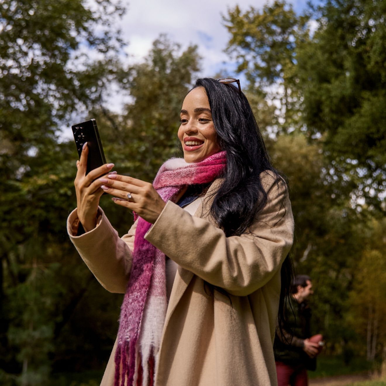 Una mujer con una bufanda a cuadros magenta y blancos está de pie al aire libre, rodeada de árboles, sonriendo a su teléfono.