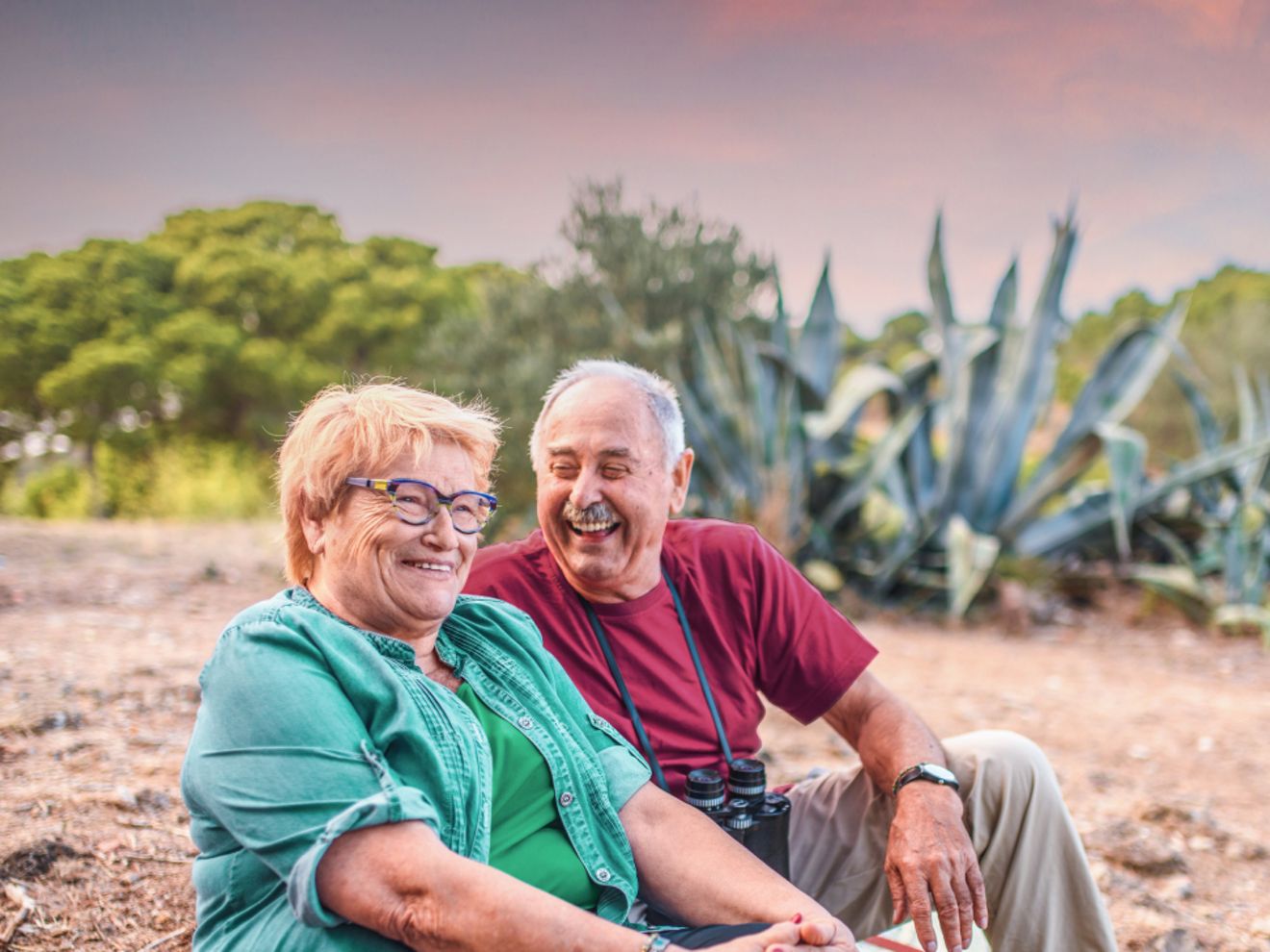An older couple are sitting and laughing while out on a hike.