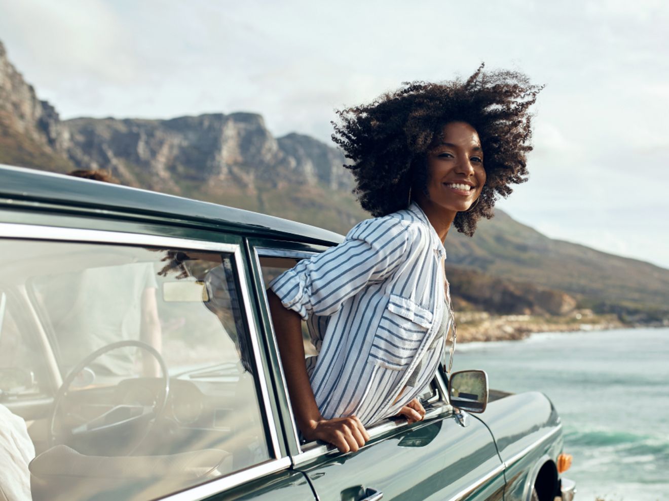 Una mujer sonriente asomada por la ventanilla de un coche estacionado en la playa.
