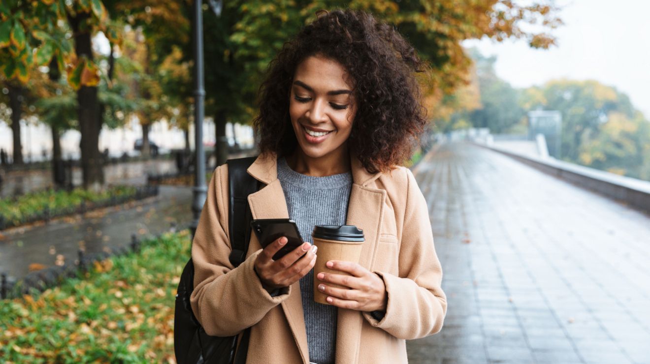 Una mujer sonríe mirando su teléfono con un café en la mano.