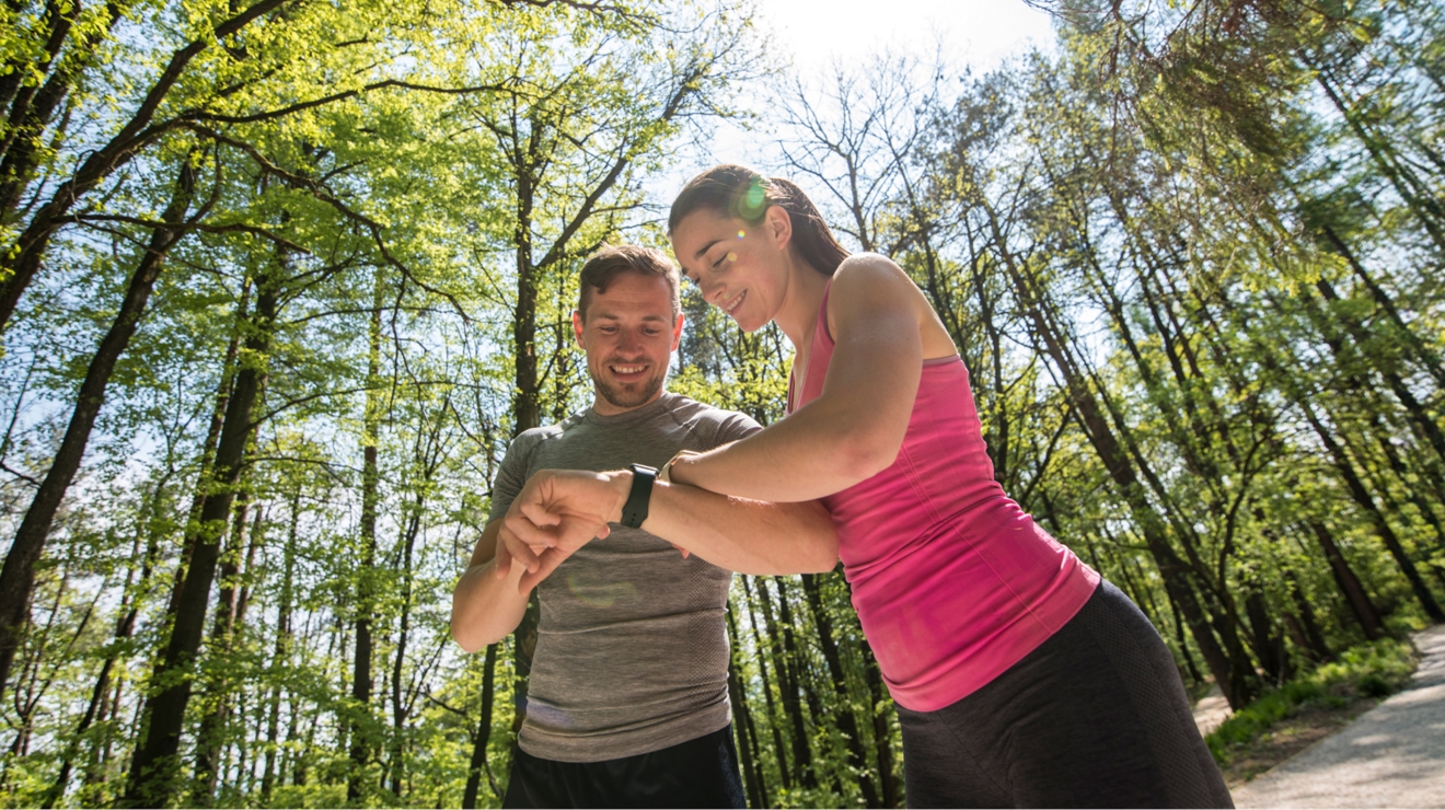 Pareja mirando sus relojes mientras hacen senderismo, rodeados de árboles.