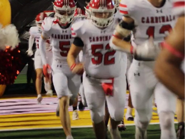 Jugadores de fútbol americano de Bridge City, vestidos con uniformes rojos y blancos, corren hacia el campo.