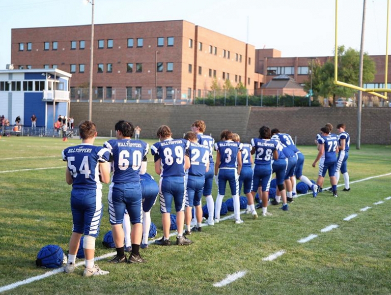 Los jugadores de fútbol de Catholic Central, vestidos con uniformes azules y blancos, permanecen fuera de la cancha durante un partido.