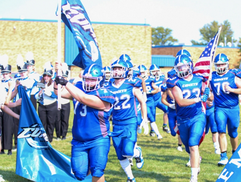 El equipo de fútbol de Coleman High School, con uniformes azules, corre hacia el campo llevando banderas.
