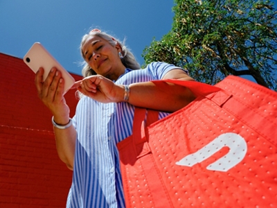 Una mujer con una camiseta a rayas azules y blancas sonríe a su teléfono mientras sostiene una gran bolsa naranja de DoorDash.