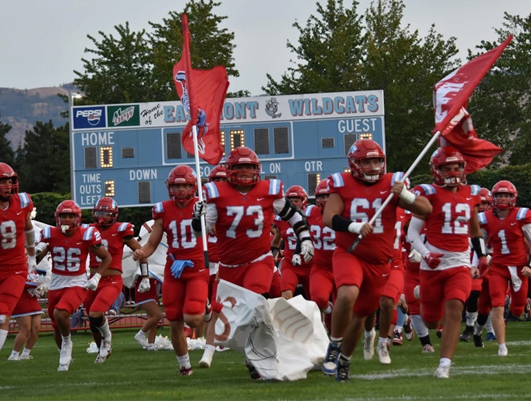 Jugadores de fútbol de Eastmont High School, vestidos con uniformes rojos y blancos, corren hacia el campo ondeando banderas.