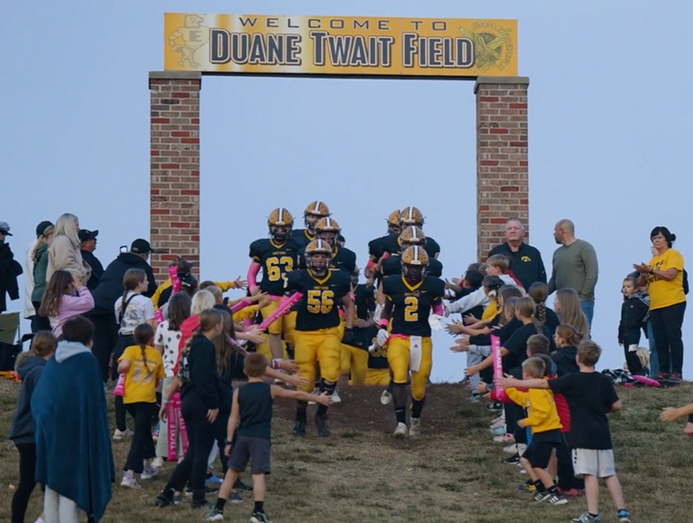 Jugadores de fútbol de Emmetsburg, vestidos con uniformes negros y amarillos, chocan los cinco con un grupo de niños mientras corren hacia el campo.