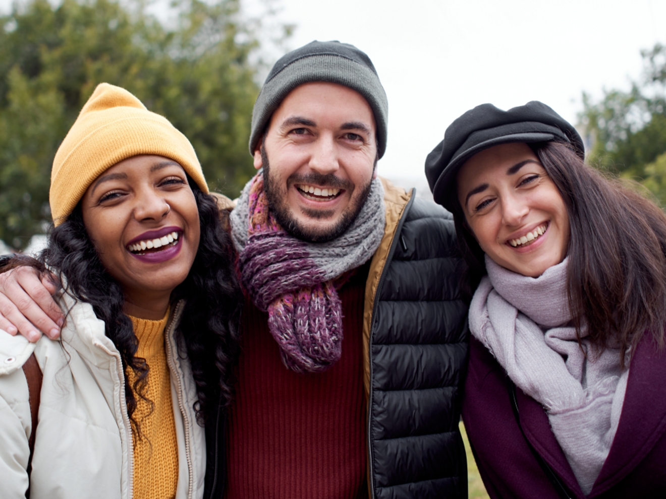 Tres amigos con abrigos y gorros de invierno sonríen abrazados al aire libre.