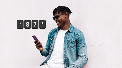 Un hombre con camiseta blanca, chaqueta de mezclilla y gafas de sol sonríe mientras usa su teléfono.