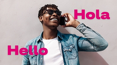 . Un hombre con camiseta blanca, chaqueta de mezclilla y gafas de sol sonríe ampliamente mientras sostiene un teléfono junto a la oreja. "Hello" y "Hola" aparecen alrededor de la persona.