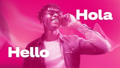 A man in a white t-shirt, denim jacket, and sunglasses smiles widely as he holds a phone to his ear. "Hello" y "Hola" aparecen alrededor de la persona.