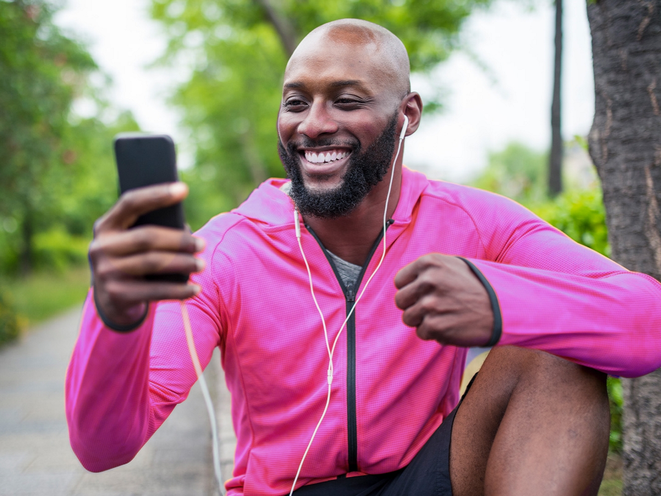 Un hombre con chaqueta magenta y audífonos con cable sentado en un parque sonriendo mientras mira su teléfono.