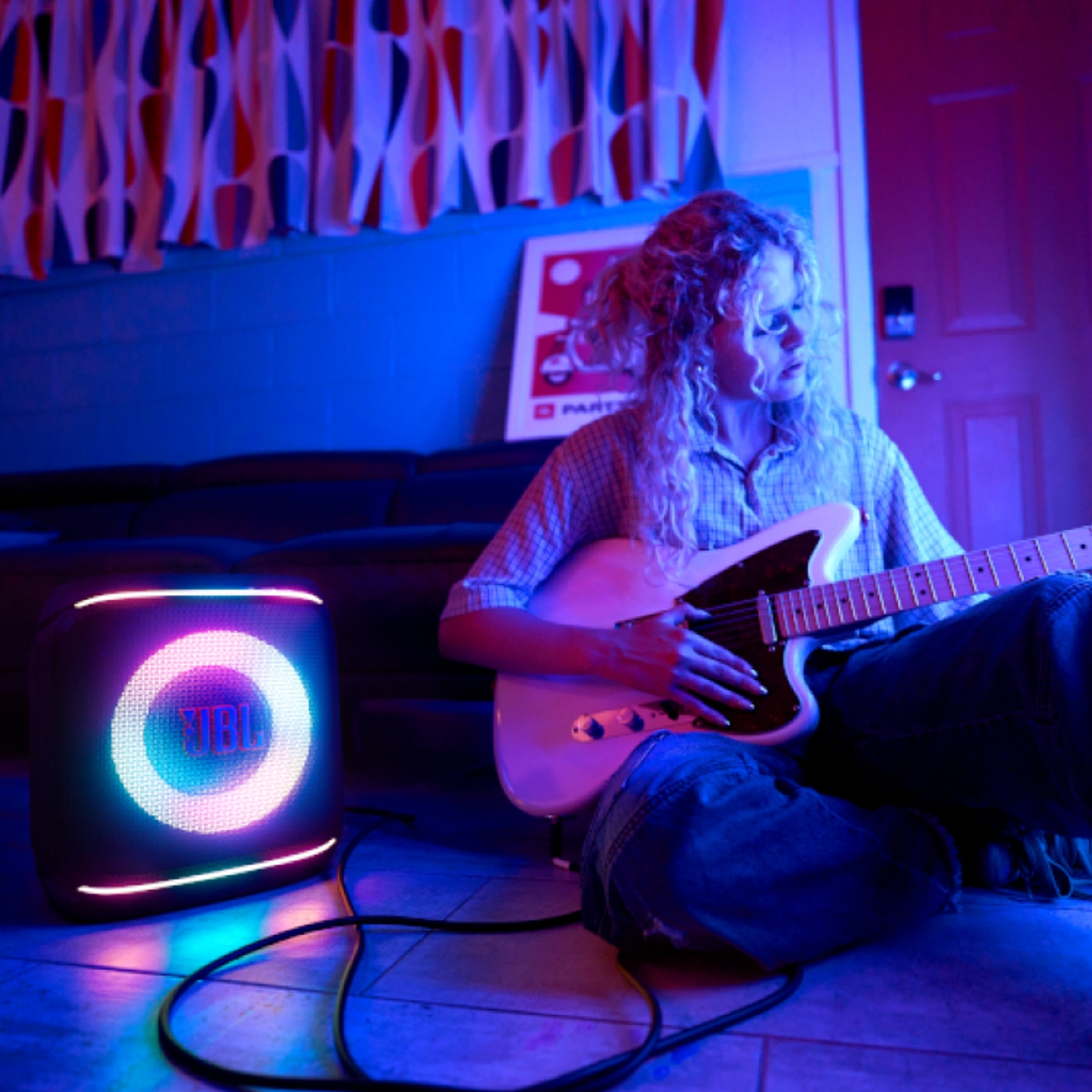 Una mujer joven tocando una guitarra eléctrica en una habitación poco iluminada mientras está sentada junto a una bocina JBL brillante.